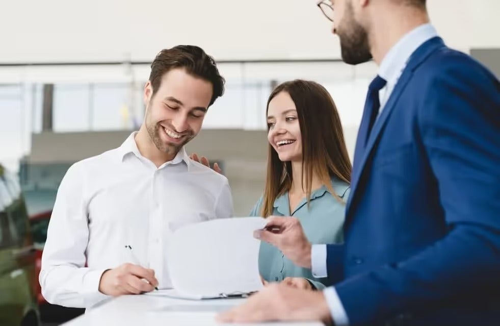 A smiling couple sign paperwork for buying a new GMC SUV with smiling salesman looking on