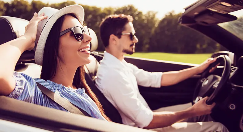 A smiling couple driving on a country road in summer; she has her hands on her hat